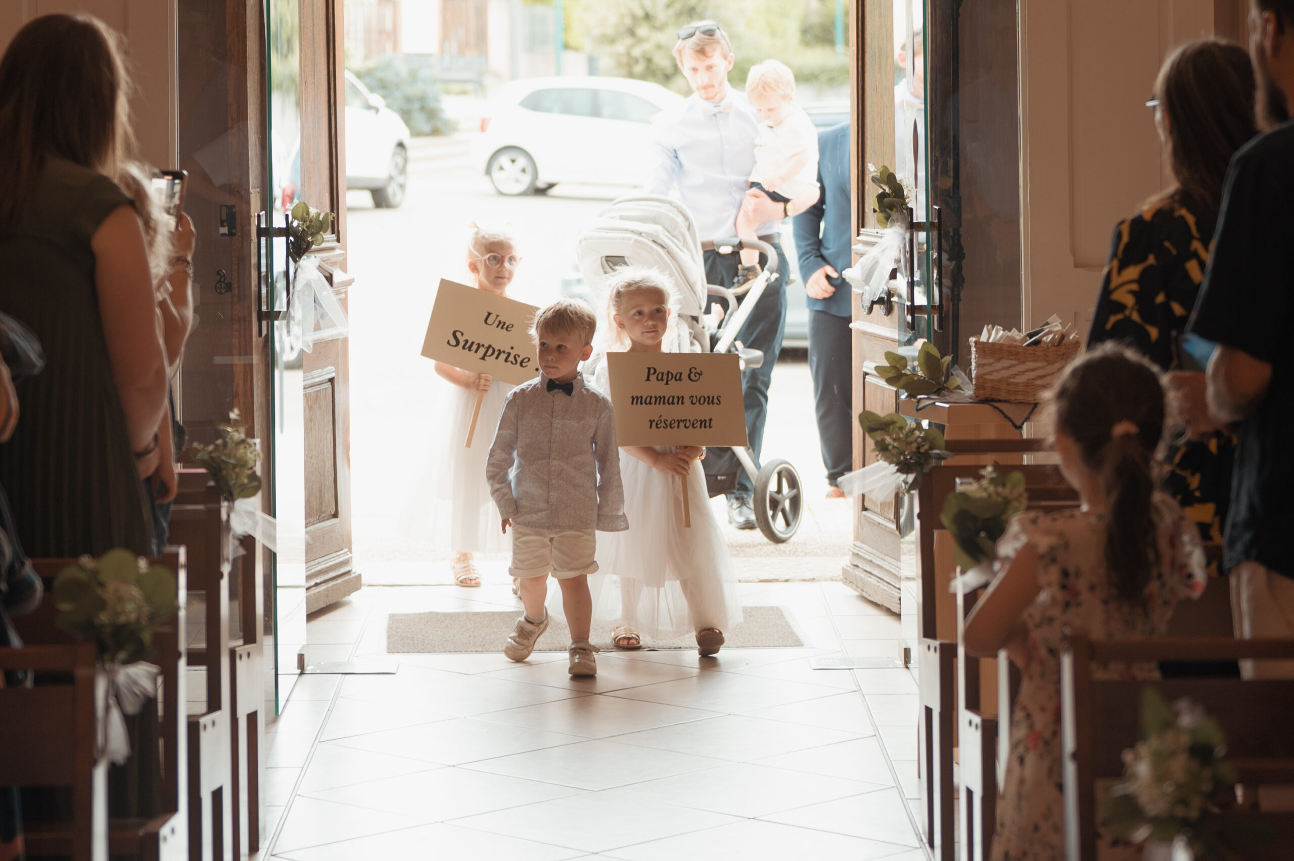 Cortège d'enfants entrant dans l'église avec des pancartes annonçant le mariage surprise de Claire et Adrien à Seyssuel