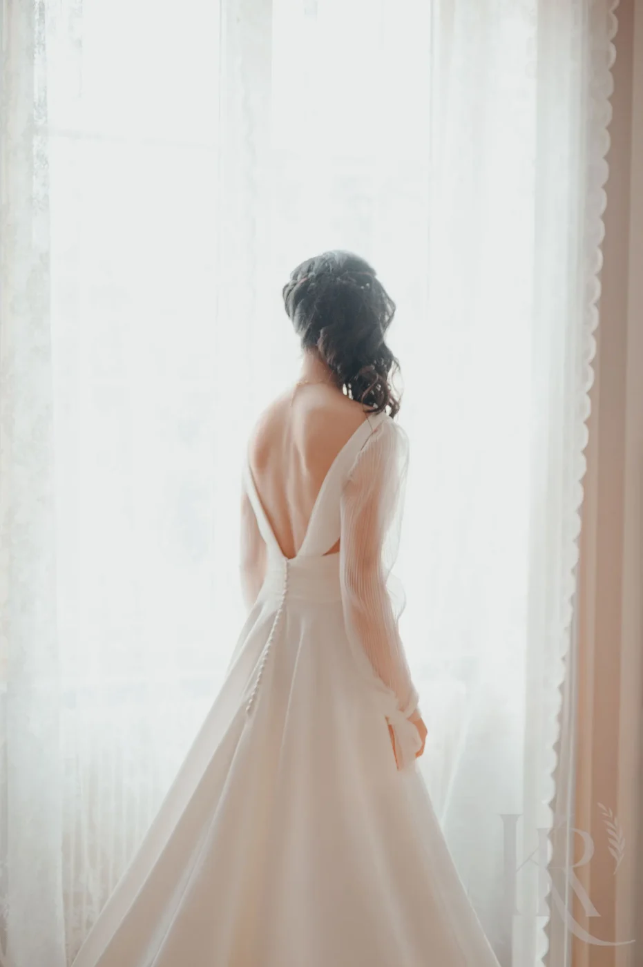 Mariée vue de dos portant robe blanche fluide dos nu devant fenêtre lumineuse