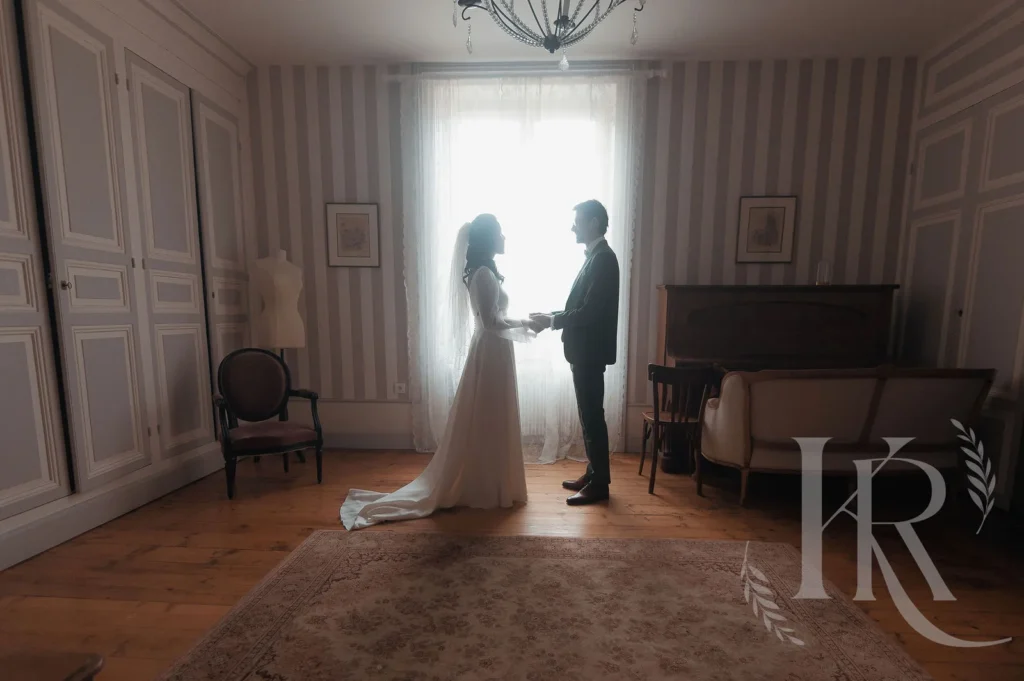 Mariée en robe blanche et marié en costume noir se tenant les mains dans un salon élégant avec fenêtre lumineuse et murs striés
