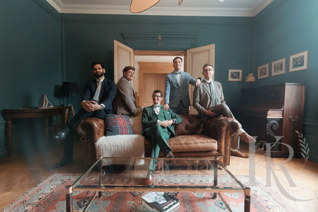 Groupe de cinq hommes en costumes élégants posant dans un salon vintage aux murs bleus turquoise avec mobilier en cuir marron.